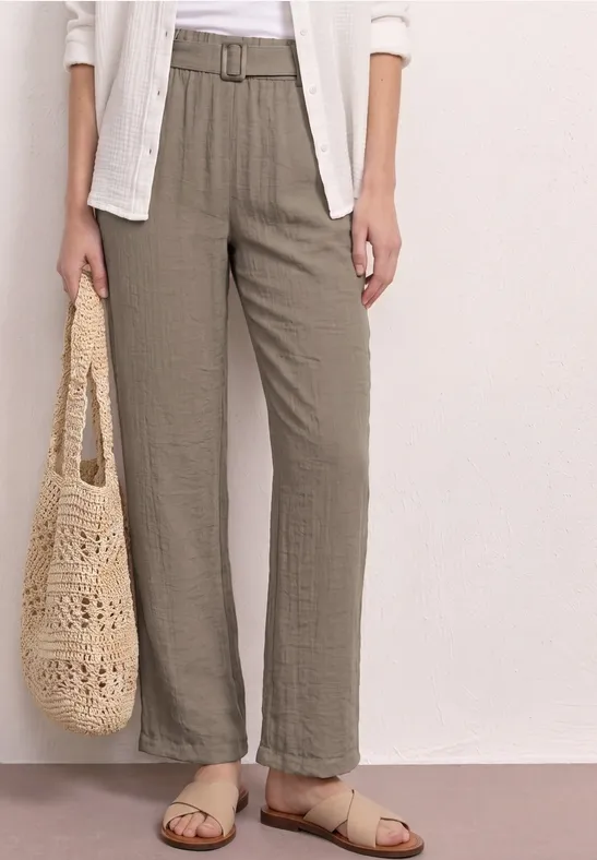 Vrouw in ruime linnen broek met riem, witte blouse, gevlochten tas en beige sandalen.