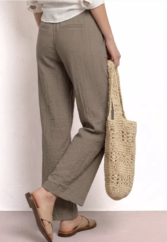Beige, wijdvallende linnen broek met platte sandalen en een gevlochten strooien tas in de hand.