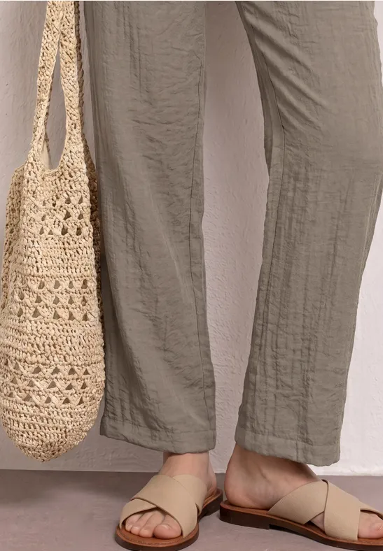 Voeten in beige riemsandalen, wijde broek en gehaakt tas.