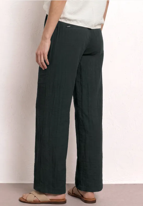 Achterzijaanzicht van een persoon met zwarte brede stoffen broek en platte sandalen tegen een lichte achtergrond.