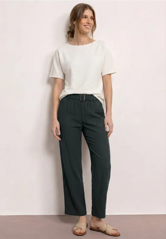 Persoon voor een lichte achtergrond, draagt een wit t-shirt, een groene broek en sandalen.