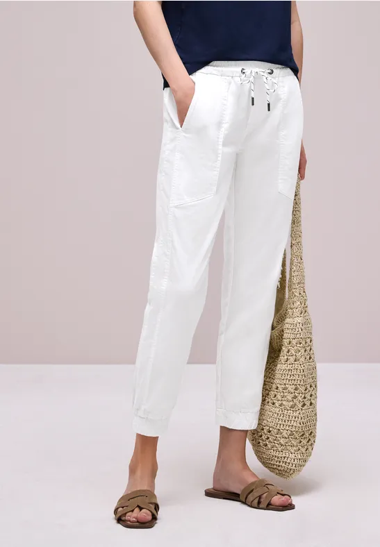 Witte linnen broek met koord en zakken, gecombineerd met sandalen en gevlochten rieten tas.