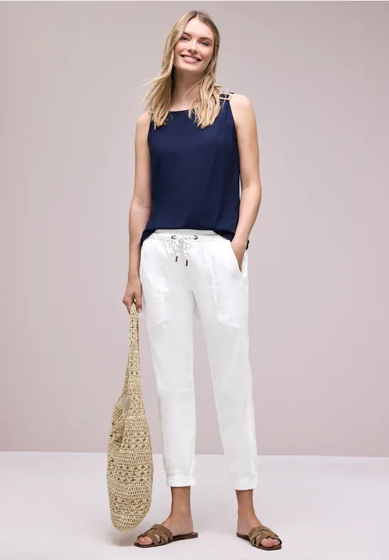 Vrouw in marineblauwe top, witte broek, sandalen en gevlochten strandtas, neutrale achtergrond.
