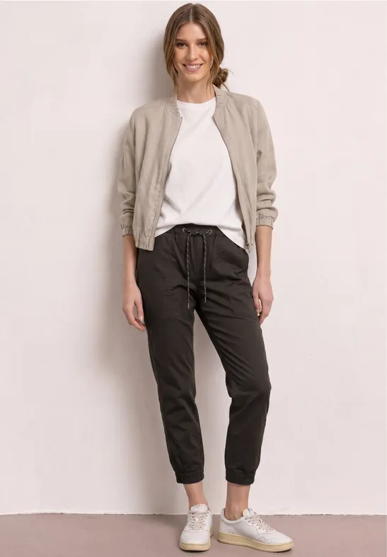 Vrouw in wit T-shirt, beige jasje, zwarte joggerbroek en witte sneakers voor een neutrale achtergrond.