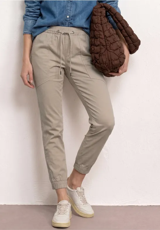 Een persoon draagt een beige joggingbroek, een denimblouse en witte sneakers, met een gewatteerde bruine tas.