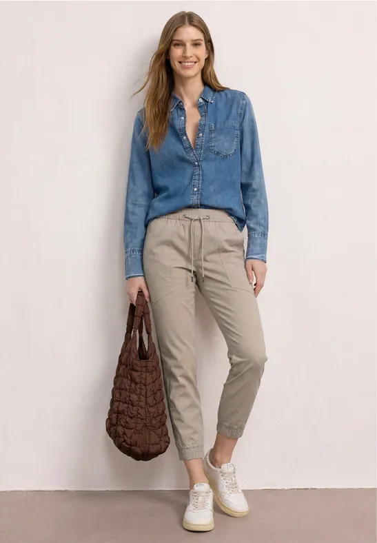 Vrouw in denim blouse, beige broek en witte sneakers, met een gevlochten tas.