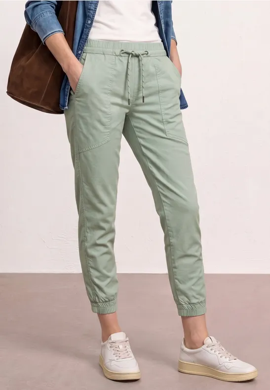 Model in groene joggingbroek met boorden, witte sneakers en spijkerhemd.