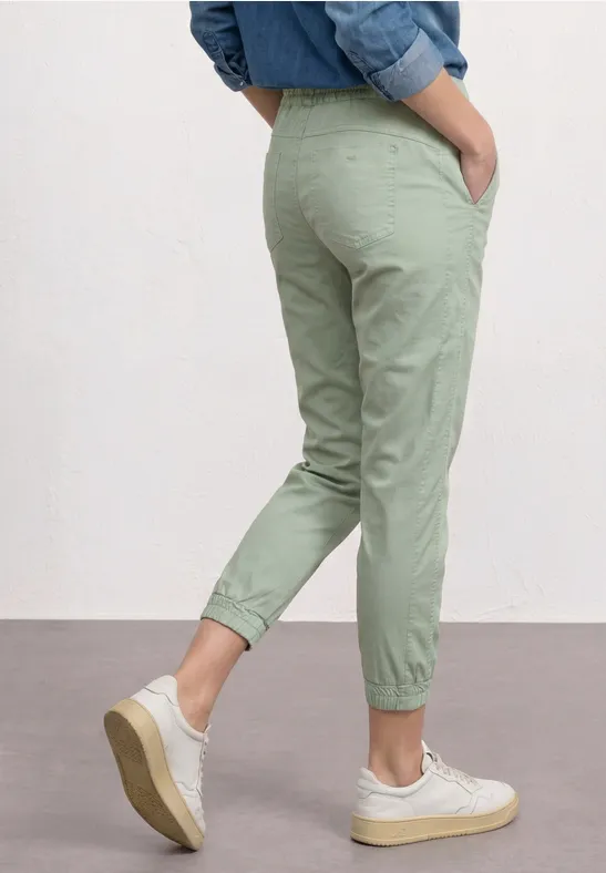 Persoon in lichtgroene, smal gesneden joggingbroek met boord en witte sneakers, zijaanzicht.