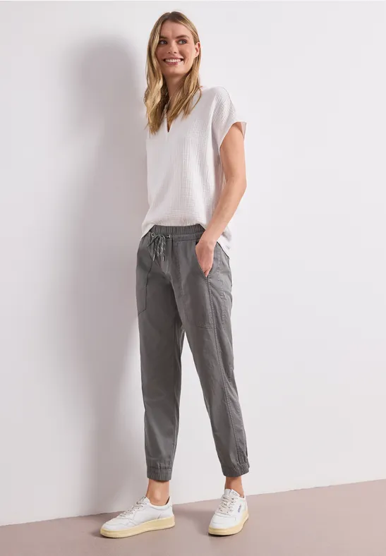 Een vrouw leunt tegen een witte muur, draagt een wit t-shirt, grijze joggingbroek en witte sneakers.