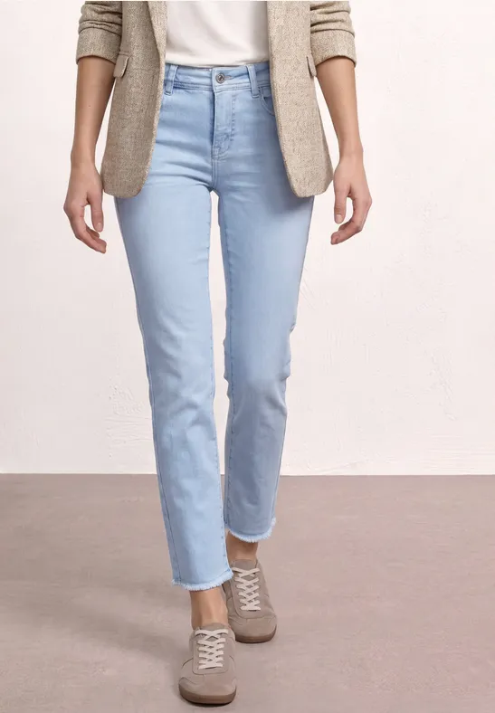 Vrouw met lichte jeans, beige vest en comfortabele sneakers tegen een neutrale achtergrond.