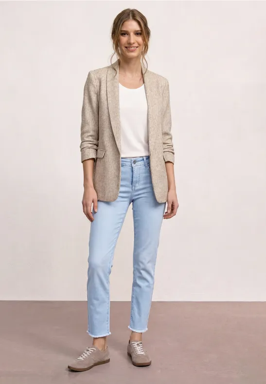 Een vrouw draagt een beige blazer, wit T-shirt, lichte spijkerbroek en grijze sneakers voor een effen achtergrond.