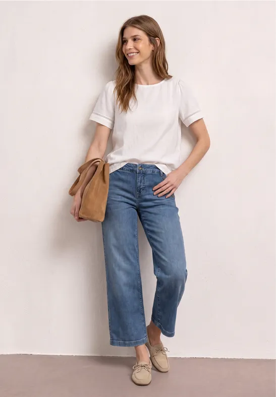Femme en t-shirt blanc, jean large court, espadrilles et sac besace beige, posant devant un mur clair.