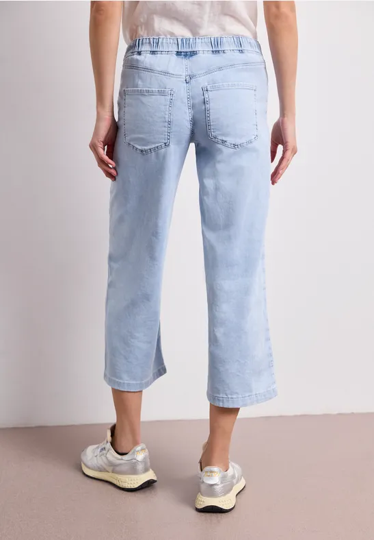 Achteraanzicht van een persoon in lichtblauwe, enkellange spijkerbroek en beige sneakers op een neutrale ondergrond.