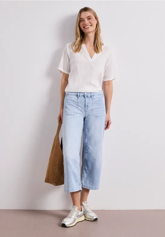 Een vrouw in een witte blouse, wijde lichtblauwe 3/4-jeans en witte sneakers, met een stoffen tas.