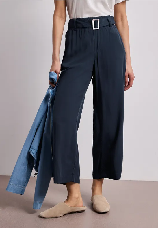 Eine Frau trägt dunkelblaue, weit geschnittene Culotte-Hosen mit Gürtel, weiße Bluse, beige Mules, hält Jeansjacke.