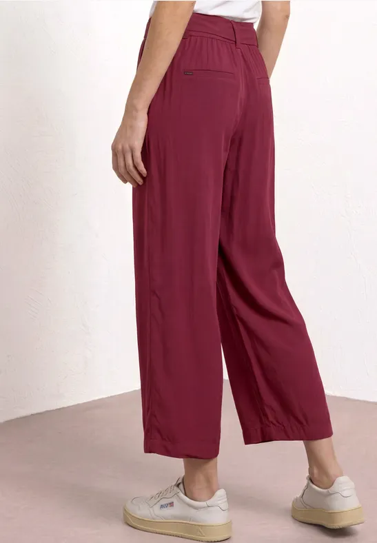 Frau trägt weite, bordeauxrote Culotte-Hose mit hoher Taille und weißen Sneakern vor hellem Hintergrund