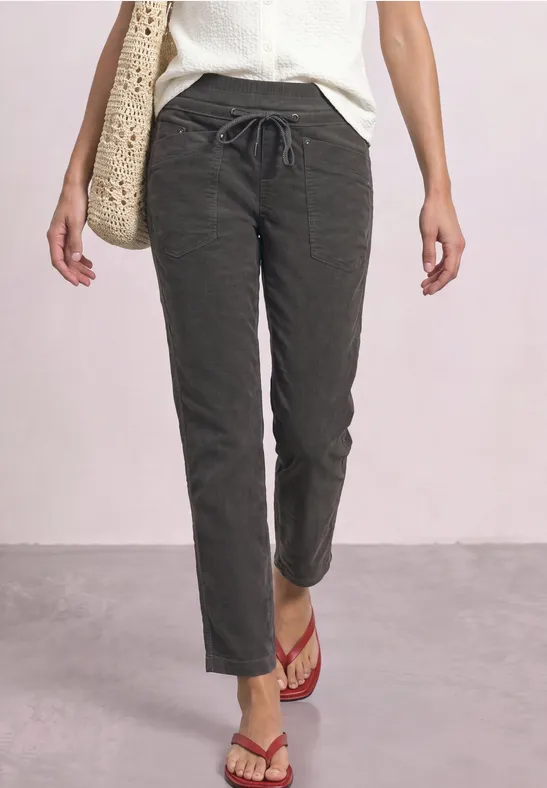 Persoon staat in donkere, casual broek, witte top, rode slippers en draagt een strooien tas.