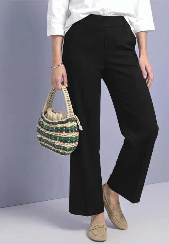 Person trägt schwarze, weite Hose, weiße Bluse, gestreifte Strohtasche und beige Slipper.