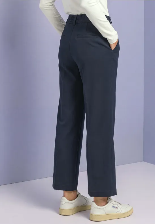 Persoon van achteren in wijde donkerblauwe broek met zakken, witte top en witte sneakers.