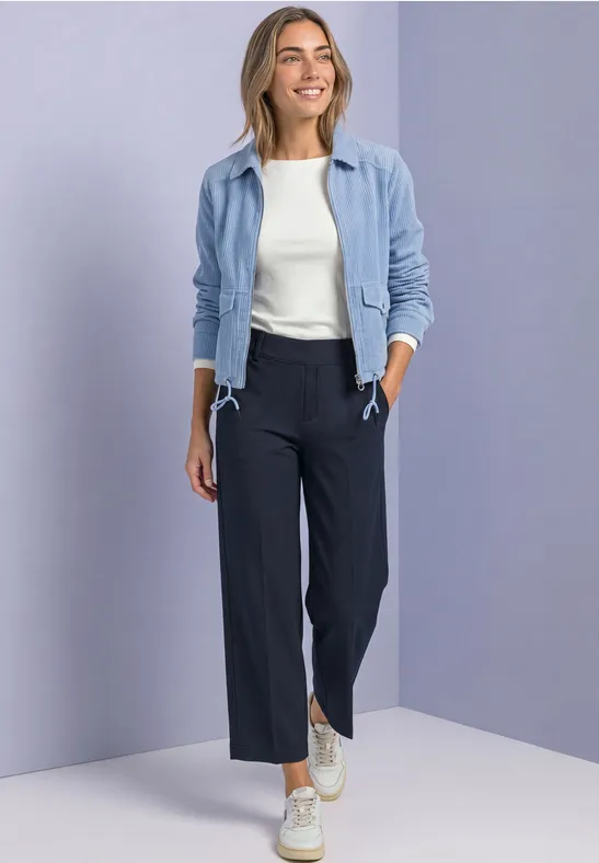Vrouw in lichtblauwe jas, wit topje, donkerblauwe wijde broek en sneakers voor neutrale achtergrond.