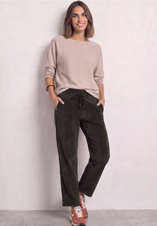Vrouw in een beige trui, donkergroene broek en oranje-witte sneakers tegen neutrale achtergrond.