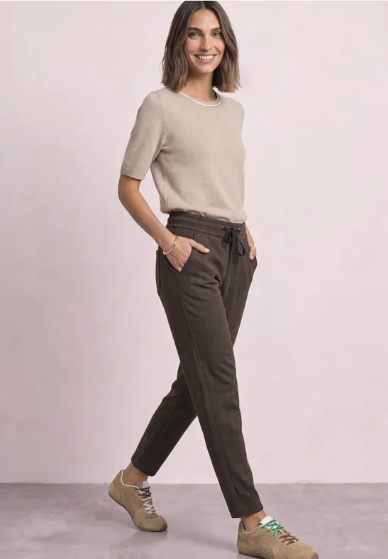 Een glimlachende vrouw in een beige T-shirt, bruine broek en beige schoenen, handen in de zakken.