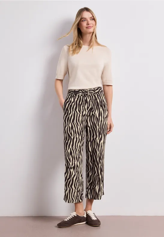 Une personne porte un t-shirt crème à manches courtes et un pantalon ample zébré, avec des chaussures marron.
