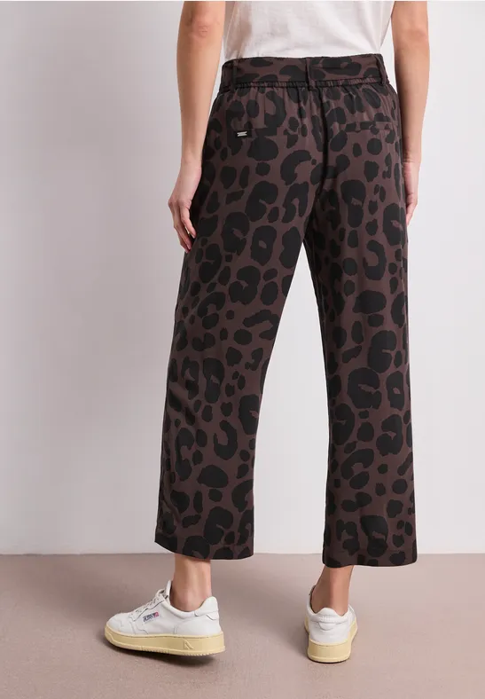 Achteraanzicht: persoon met witte sneakers, wijde broek met bruin-zwart luipaardprint en wit T-shirt.