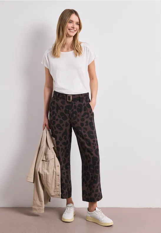Vrouw met wit T-shirt, wijde broek met print en lichte sneakers, houdt een beige jas vast.