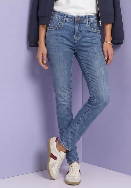 Personne en jean bleu et baskets blanches devant un mur violet clair.