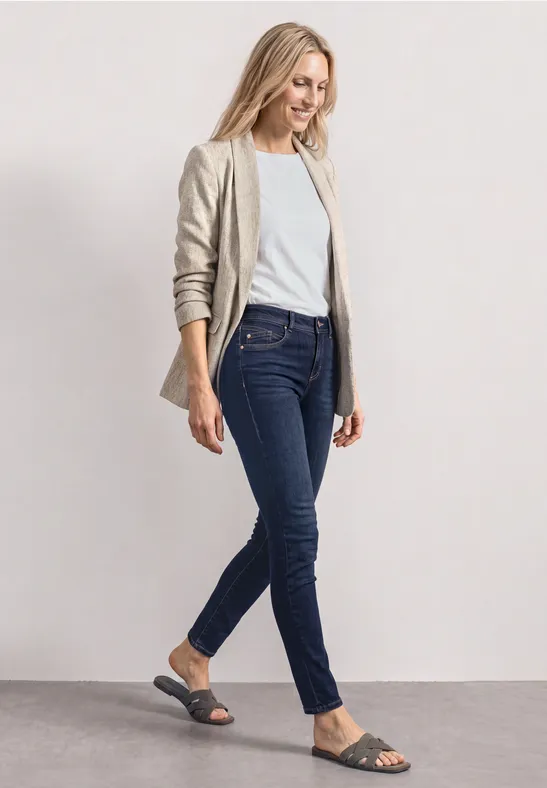 Une femme en blazer beige, t-shirt blanc, jean bleu foncé et sandales noires marche.