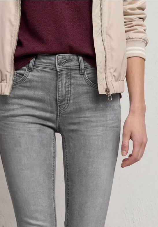 Eine Person trägt graue Jeans, einen burgunderroten Pullover und eine helle Jacke, Sicht auf Taille und Hände.