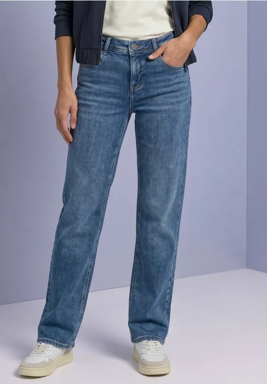 Une personne porte un jean droit bleu et des baskets blanches devant un fond neutre.