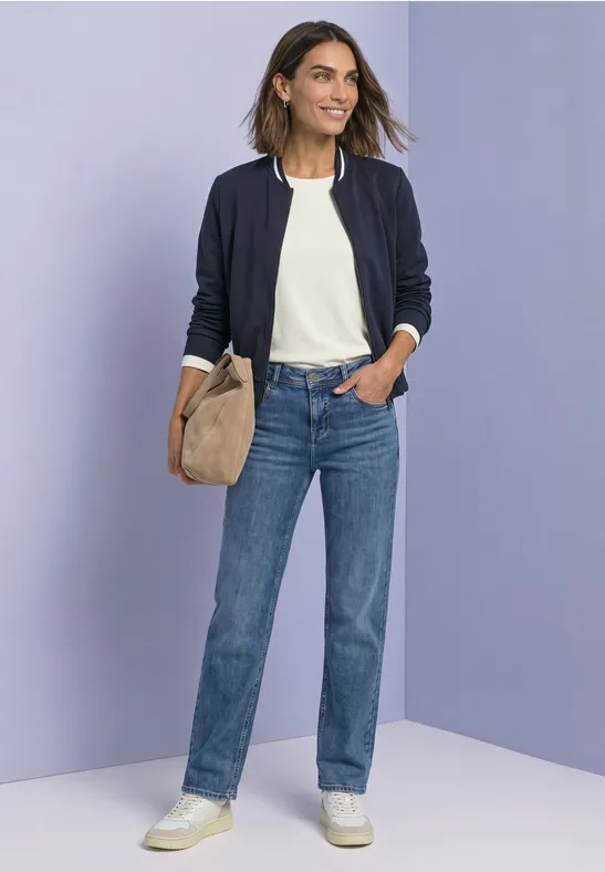Femme en t-shirt blanc, jean bleu, veste foncée et baskets blanches, sac beige, fond violet.