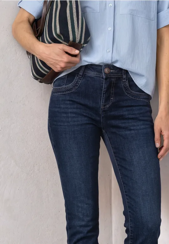 Person in hellblauem Kurzarmhemd und dunkelblauen Jeans hält gestreifte Tasche unter dem Arm vor neutralem Hintergrund.