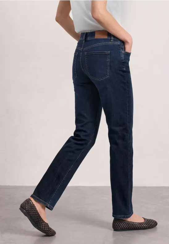 Vue de profil d'une personne en jean bleu foncé et mocassins plats, marchant devant un fond clair.