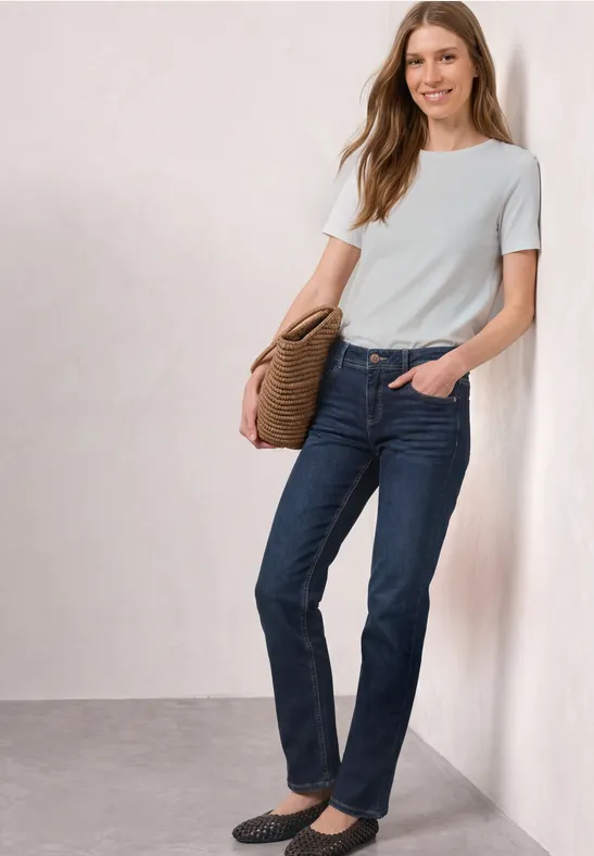 Femme appuyée contre un mur, en t-shirt blanc, jean bleu, mocassins noirs et sac marron.