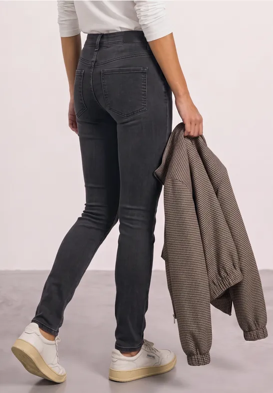 Vue de dos d'une personne en skinny noir, baskets blanches aux pieds et veste kaki tenue à la main.