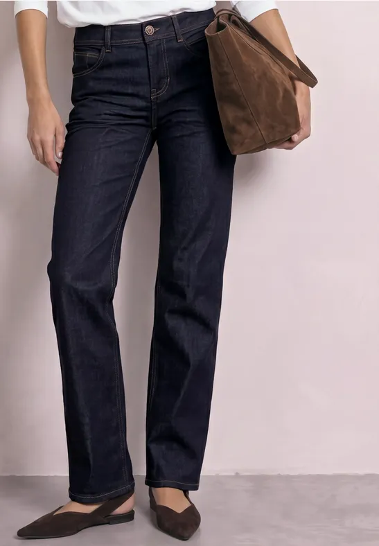 Persoon draagt rechte, donkerblauwe jeans, zwarte ballerina’s en houdt een bruine tas vast.
