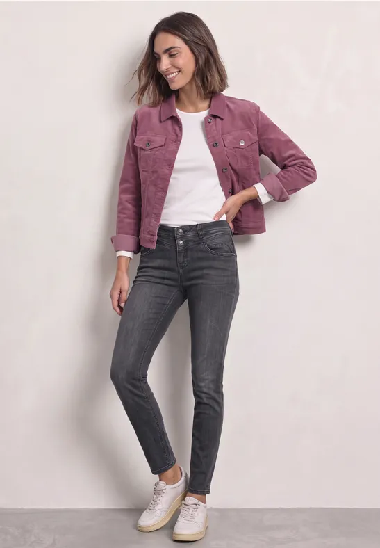 Jeune femme porte une veste rose, t-shirt blanc, jean gris foncé et baskets blanches devant un fond clair.