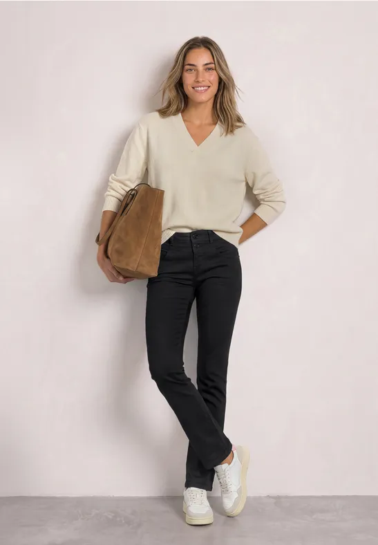 Une femme souriante porte un pull crème, un pantalon noir, des baskets blanches et tient un sac marron.