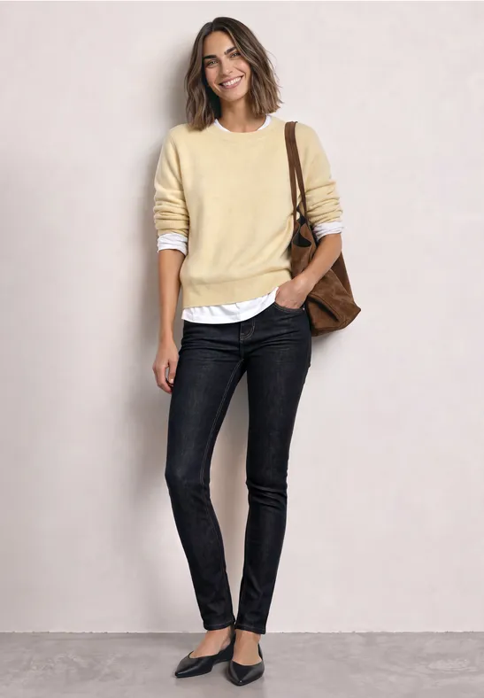 Eine Frau trägt einen hellgelben Pullover über weißem Hemd, schwarze Jeans, spitze Ballerinas und eine braune Schultertasche.