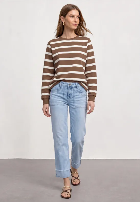 Vrouw in bruin-wit gestreepte trui, blauwe jeans en sandalen voor een lichte achtergrond.