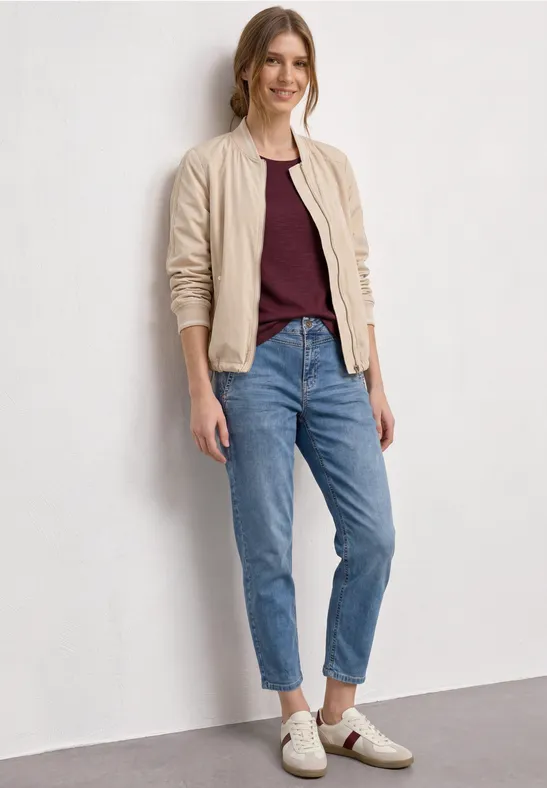 Persoon draagt beige bomberjack, bordeaux T-shirt, blauwe jeans en witte sneakers.