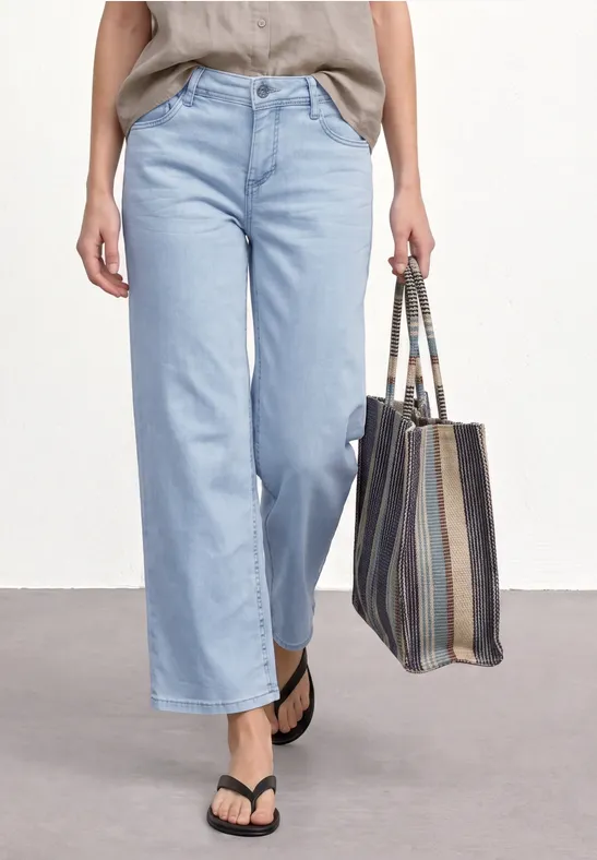 Une personne porte un jean large bleu clair et des tongs noires, tenant un sac rayé en tissu.