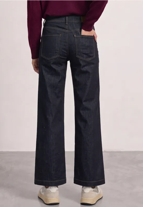 Vue arrière d’une personne portant un jean large et sombre avec des baskets sur un fond neutre.