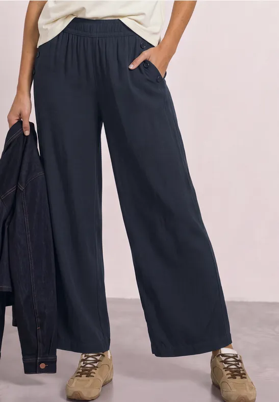 Vrouw in wijde donkerblauwe broek met zakken, wit topje en beige sneakers, houdt een jas vast.