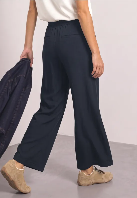 Persoon draagt wijde zwarte broek en beige schoenen, houdt een zwart jasje in de hand.
