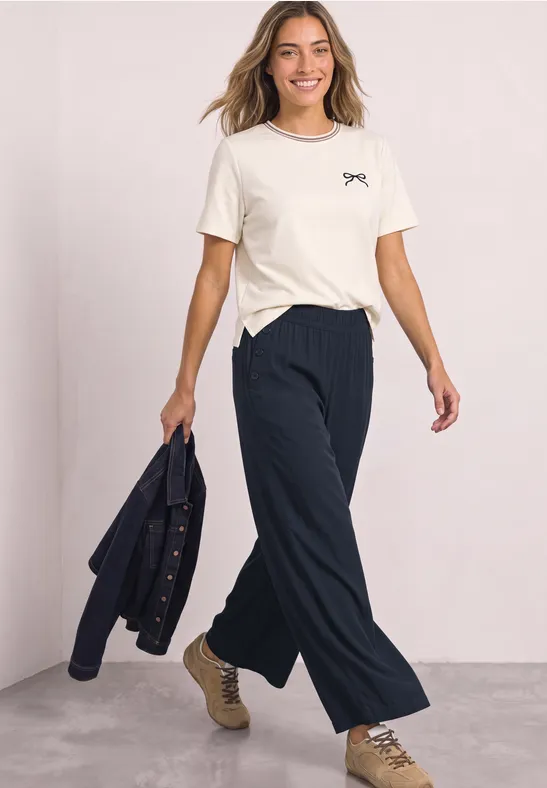 Een glimlachende vrouw draagt een wit T-shirt, losse zwarte broek en houdt een jas in haar hand.