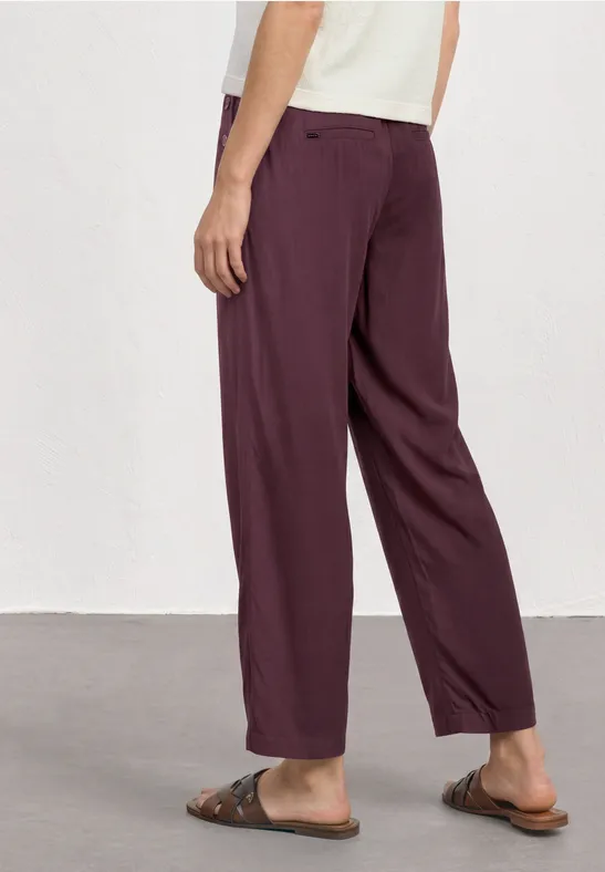 Een persoon draagt wijde bordeauxrode broek met zakken en bruine sandalen, voor een witte achtergrond.